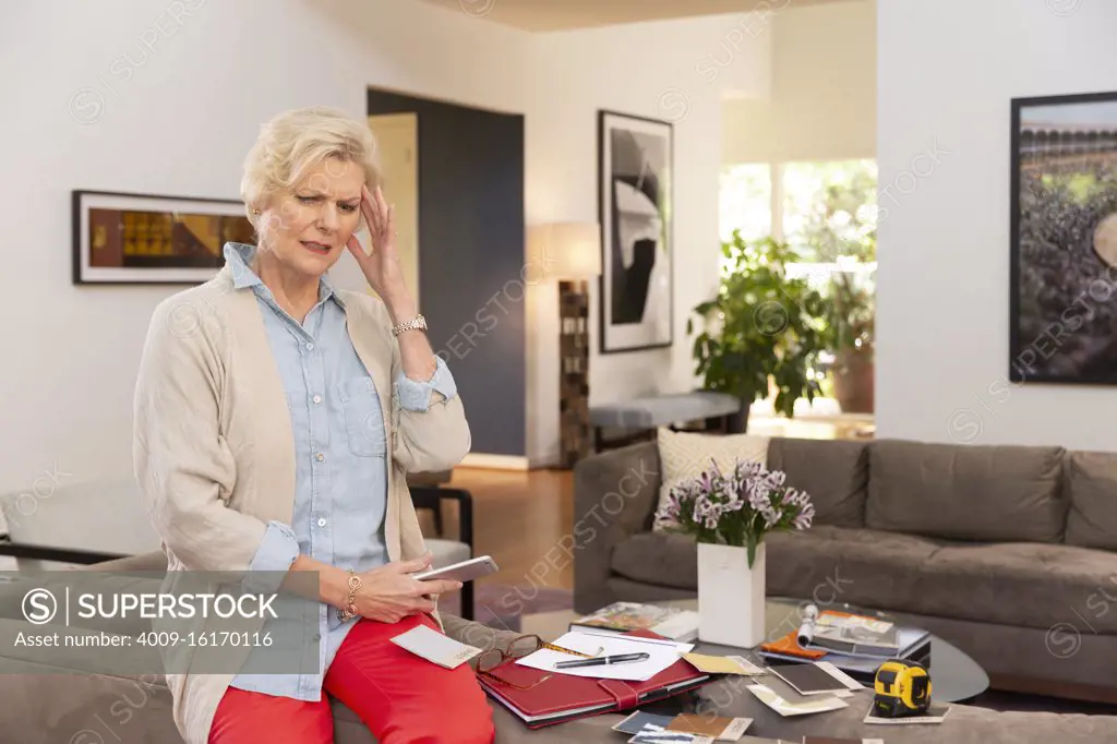 Mature Caucasian female designer working from home, sitting on back of couch with fabric swatches and notes laid out around her, Having headache while using cell phone to get medical advice and treatment 
