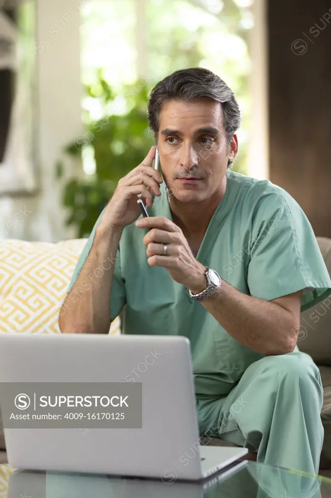 Hispanic Male doctor practicing tele-medicine from his home, using cell phone and laptop computer, Listening to patient on video call 