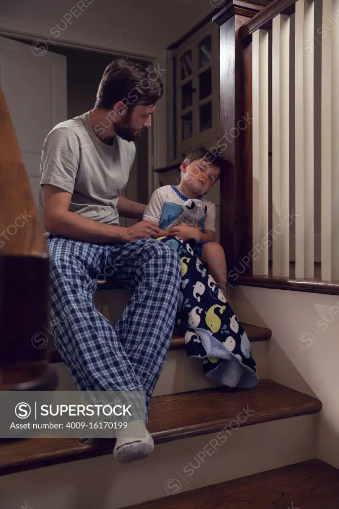 Father and son sitting at top of stairs in home getting ready for bedtime, dad checking on his son who is feeling ill 