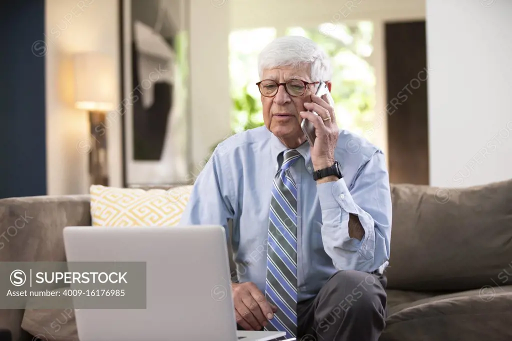 Mature Caucasian Male doctor practicing tele-medicine from his home, using cell phone and laptop computer, Listening to patient on video call 