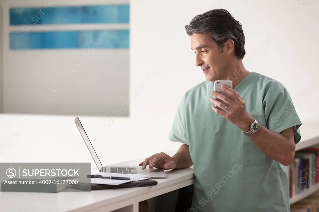 Hispanic Male doctor in his home practicing Telemedicine, talking on cell phone and using laptop computer 