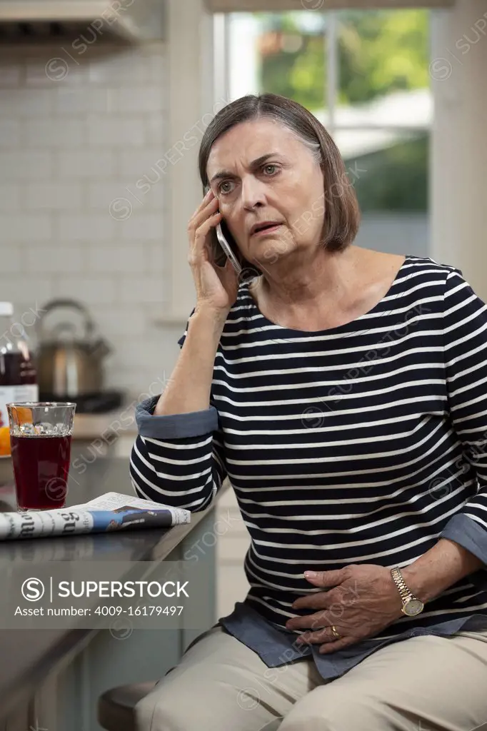 Senior Caucasian woman in her kitchen describing her abdominal pain to doctor on the phone