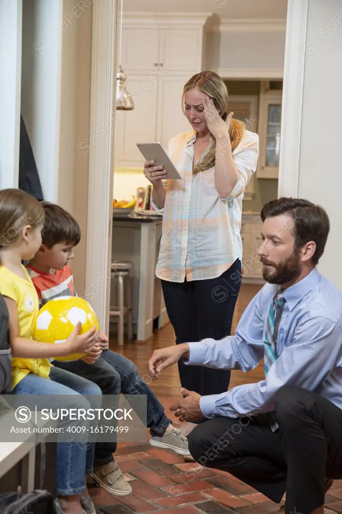 Family in the morning getting, dad getting kids shoes on in mudroom before taking off for school, Mom talking with doctor on iPad describing her symptoms 