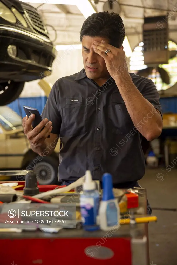 Hispanic car mechanic working in auto shop, having migraine headache, using mobile phone to check in with tele-medicine doctor 