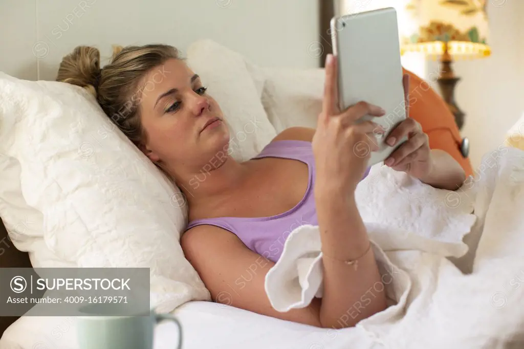 Young woman in bed debating getting up, using tablet to check in with doctor 