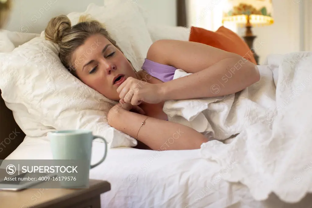 Coughing young woman in her bed debating getting out of bed, smartphone and coffee mug sitting on bedside table 
