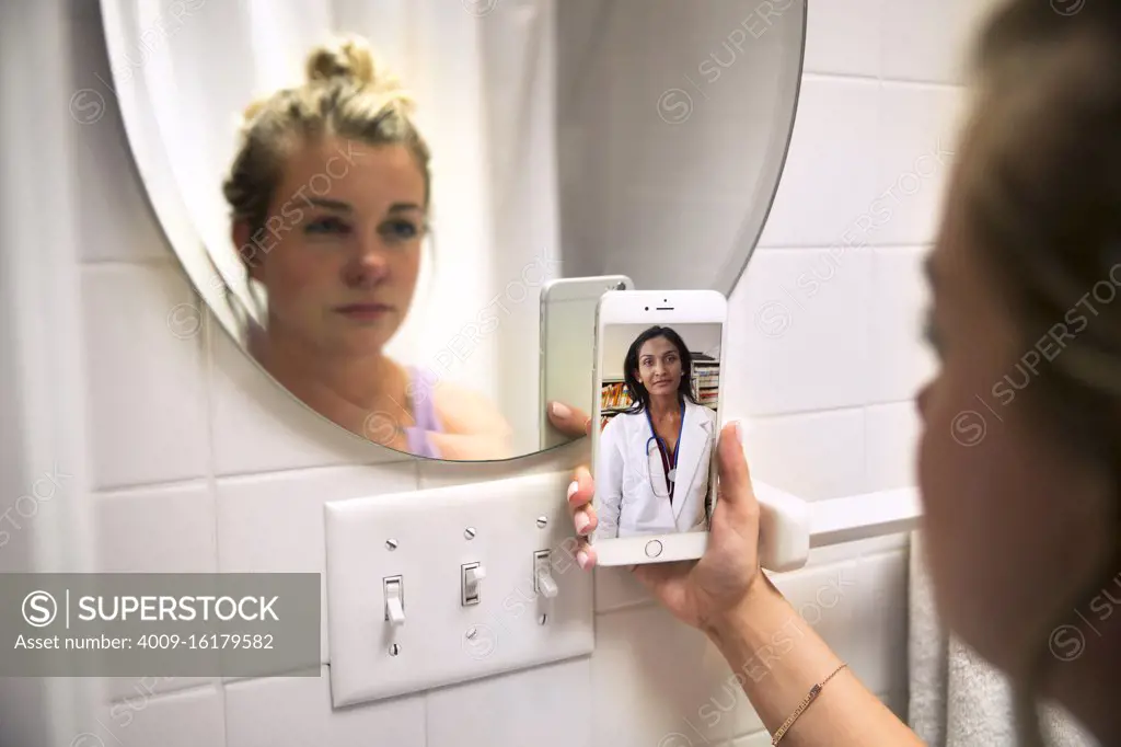 Young woman in her bathroom looking at herself in mirror while talking to doctor on video call