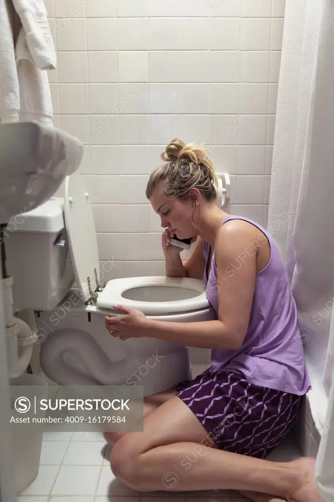 Young woman on the floor in her bathroom feeling sick next to toilet, talking to doctor on cellphone 