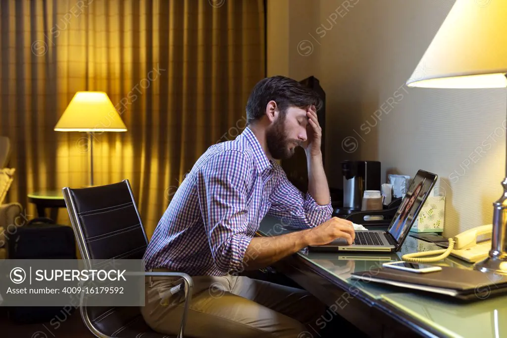 Caucasian man working late in a hotel room, feeling ill so he is having telemedicine meeting with doctor on laptop 