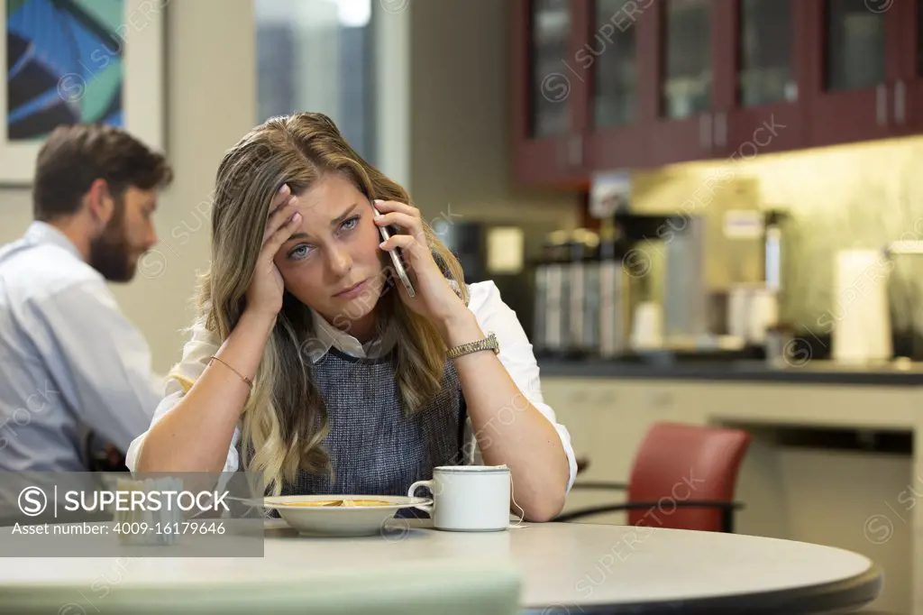 Young Caucasian woman with headache talking on cell phone while sitting in break area of office with lunch of soup and Hot Tea