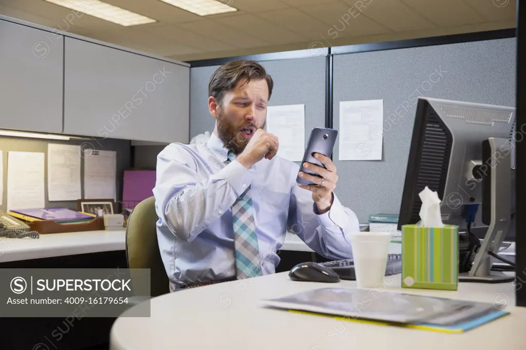 coughing young Caucasian man working in cubicle at office, fighting off a cold with tissues and hot tea, checking cell phone 