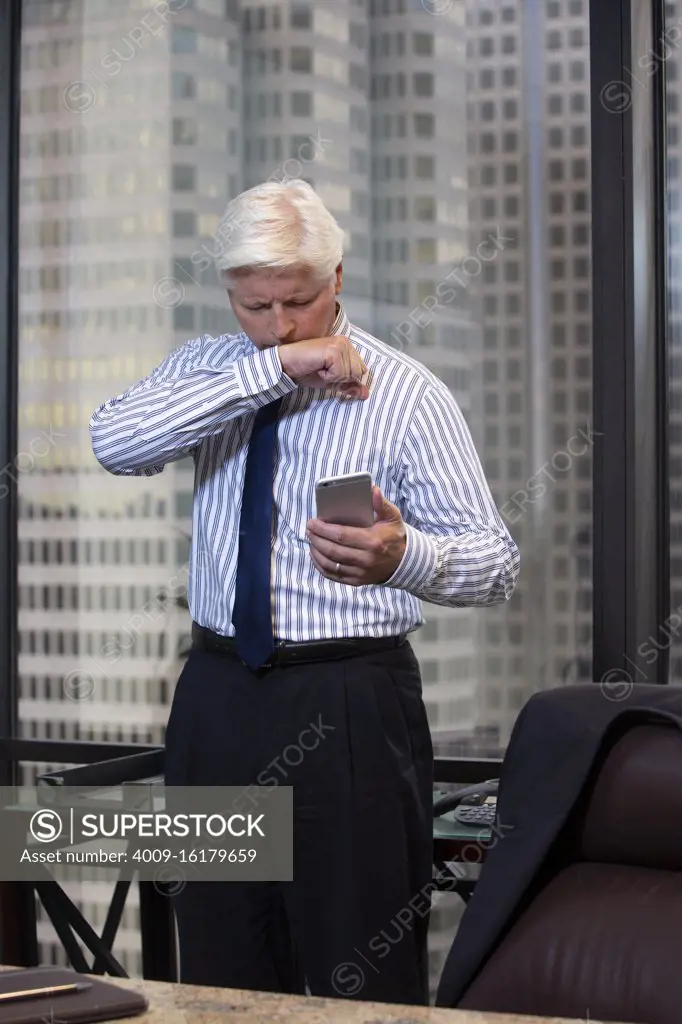 Coughing mature Caucasian man fighting off a cold in office of high-rise building downtown, looking at smartphone having a tele-medicine video call with doctor 