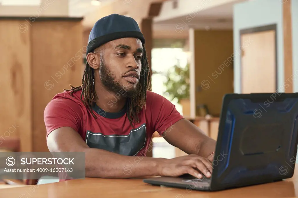 Portrait of young ethnic man wearing red t shirt and knit hat , sitting at bar in kitchen of  downtown loft  with laptop computer 