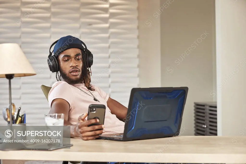 portrait young ethnic man sitting at desk with laptop computer wearing large over ear headphones looking at smartphone 