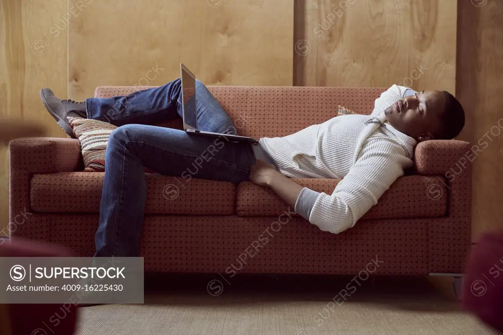 Young man taking a nap on loveseat with his computer on his lap
