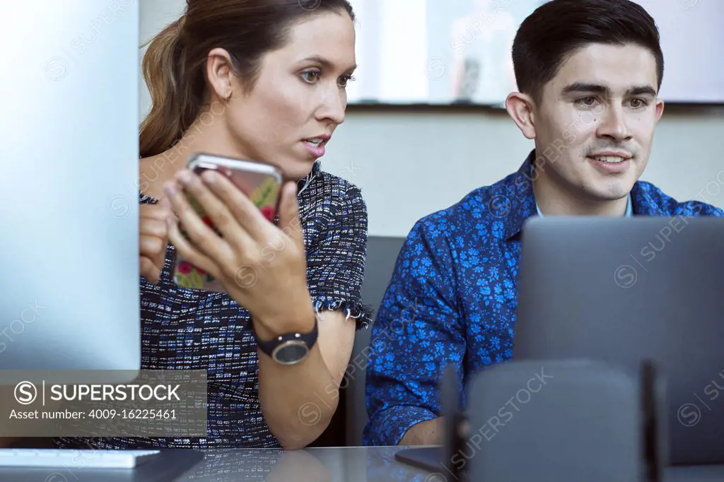Young man and woman co-workers gathered around laptop computer looking at screen, woman checking smart phone 
