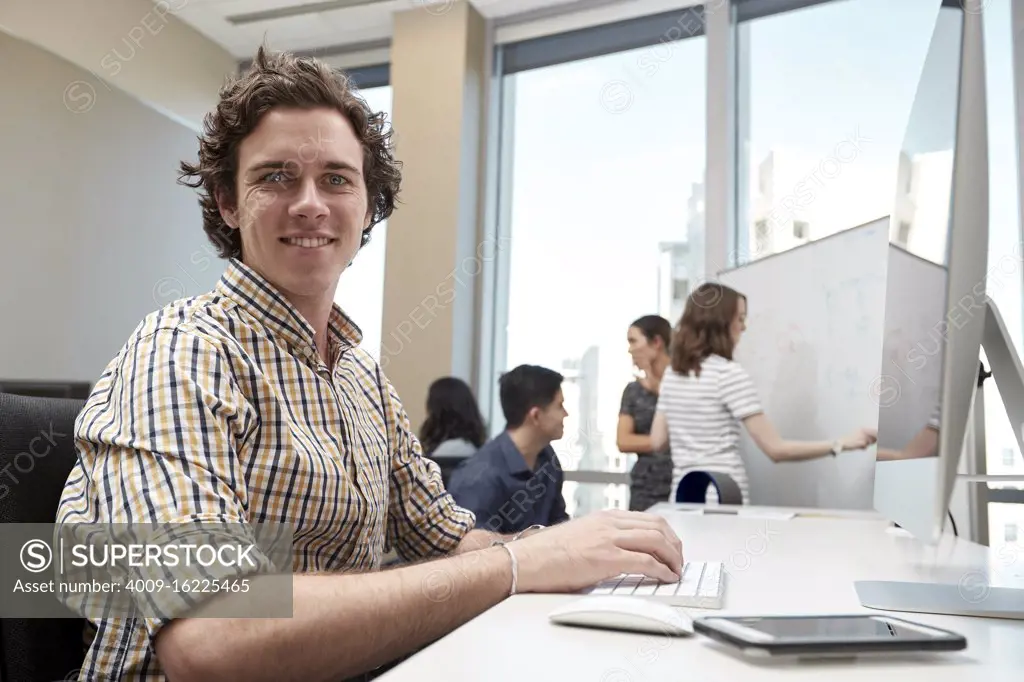 Young man using computer in office looking towards camera, co-workers having discussion in background 