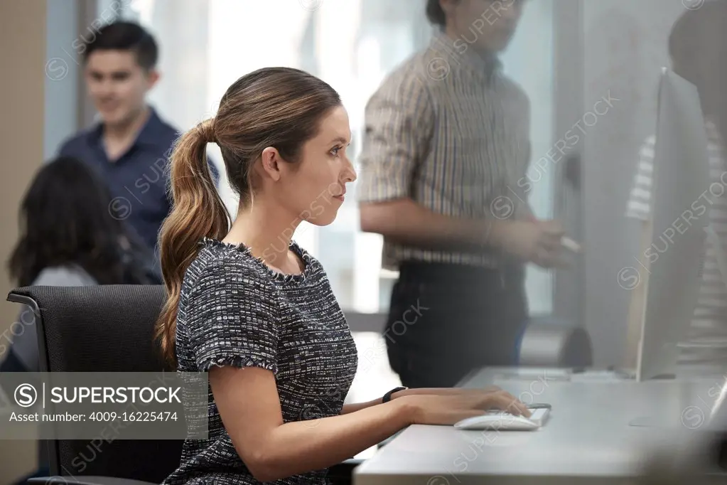 Young woman using computer in office typing on keyboard, co-workers having discussion around dry erase board in background 
