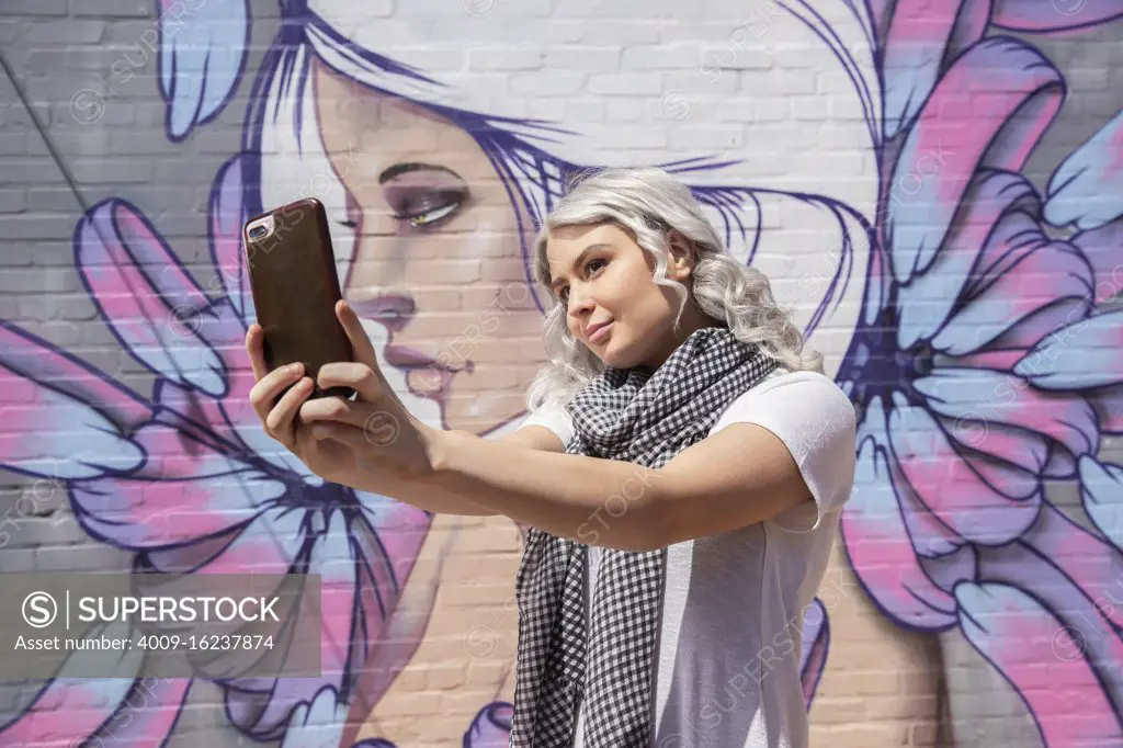 Young woman in front of graffitied wall using mobile phone to take selfie 