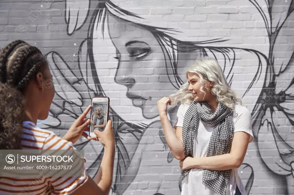 Two young women in front of graffitied wall using mobile phone to take portrait 