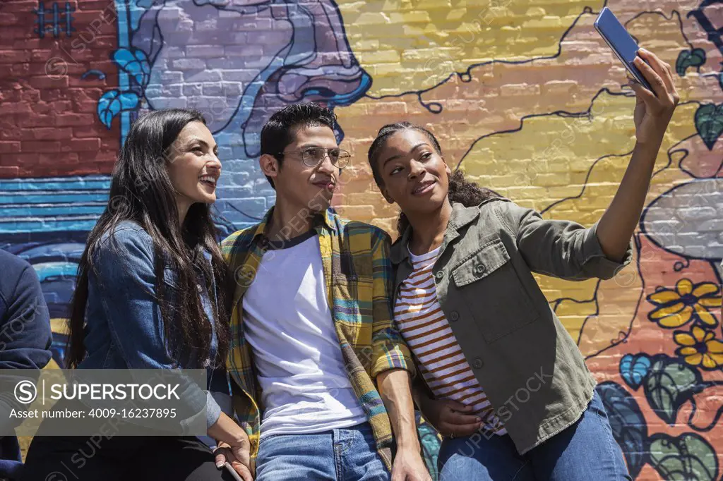 Group of young co-workers hanging out in front of graffitied wall using mobile phone to take selfie 