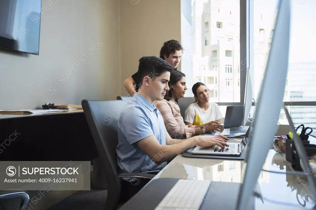 Young man using laptop computer in office conference room, co-workers having discussion in background 