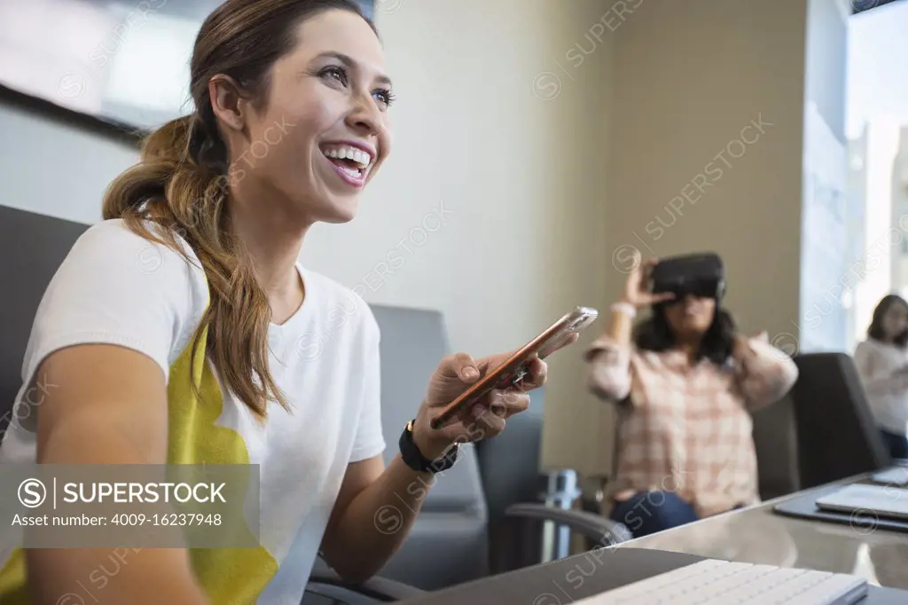 Young woman in conference room working on computer checking mobile phone, with co-worker in background wearing a Virtual reality headset 