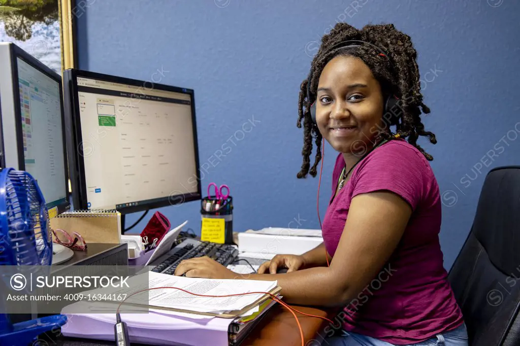 A portrait of an African American woman at her call center desk and her headset on, working from home, looking into camera smiling.