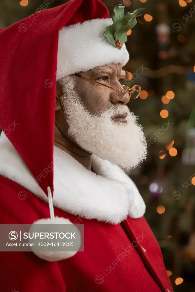 An African American man dressed as Santa Claus sitting in front of a Christmas tree turned profile, looking off camera.