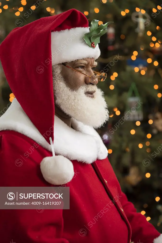 An African American man dressed as Santa Claus sitting in front of a Christmas tree turned profile, looking off camera.