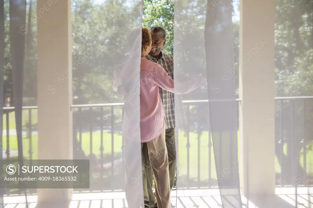 Older couple dancing on porch overlooking  green lawn , seen through sheer curtains 
