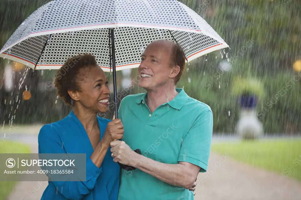 Older couple walking through park with umbrella in the rain, lovingly looking at one another  