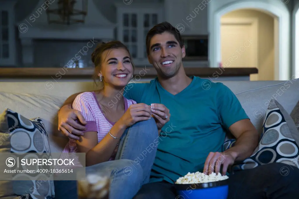 Young couple sitting on couch watching a movie at home with a bowl of popcorn 