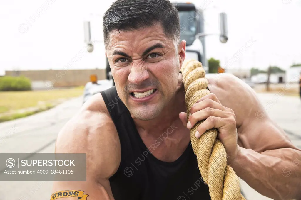 Muscular Hispanic man wearing tank top with Strong” tattoo on his bicep, Straining to pull semi truck with large tow rope 
