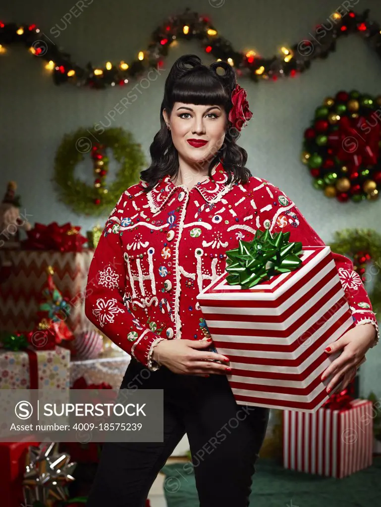 Portrait of woman with a retro hair style, in an "ugly" Christmas sweater holding a present looking at the camera.