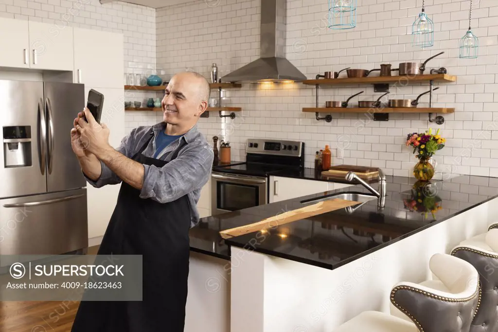 Man in kitchen wearing apron having a video call on his phone in kitchen. 