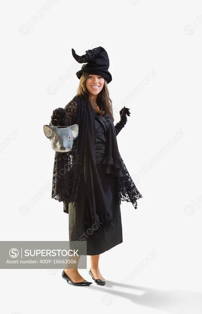 Full length shot of a Hispanic woman dressed as a witch for Halloween holding a mouse head trick or treat bucket dancing, against a white background