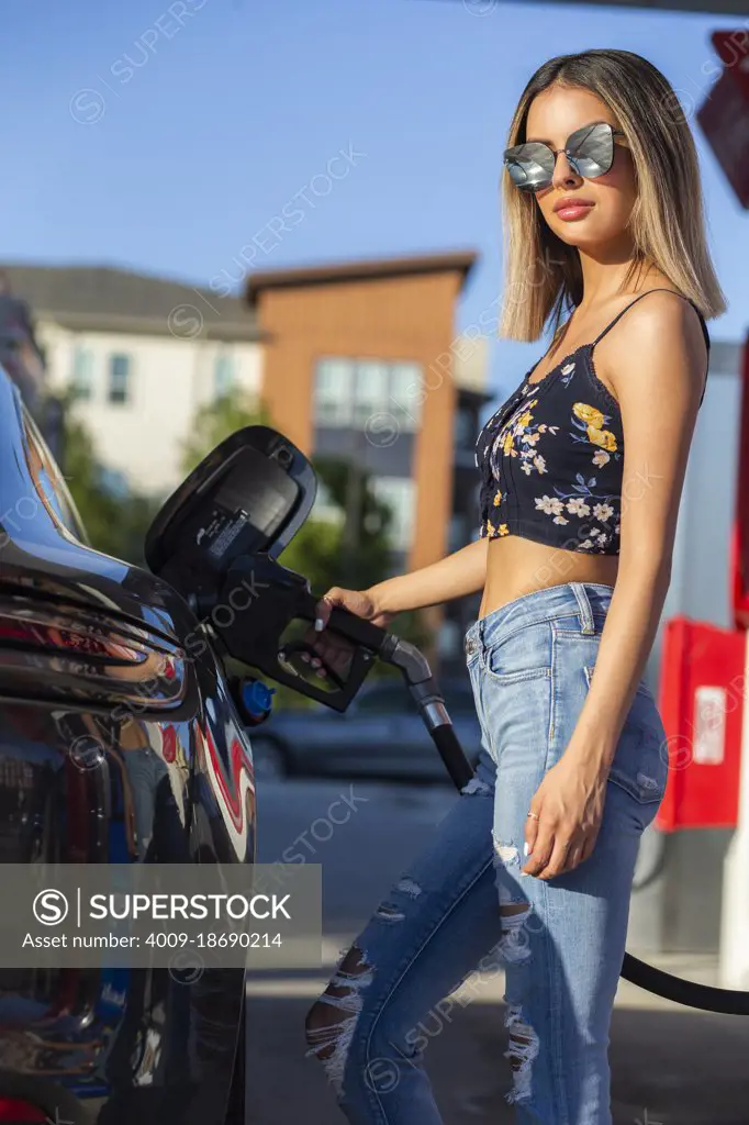 Confident young Hispanic girl standing at gas station, pumping gas into her luxury car. 