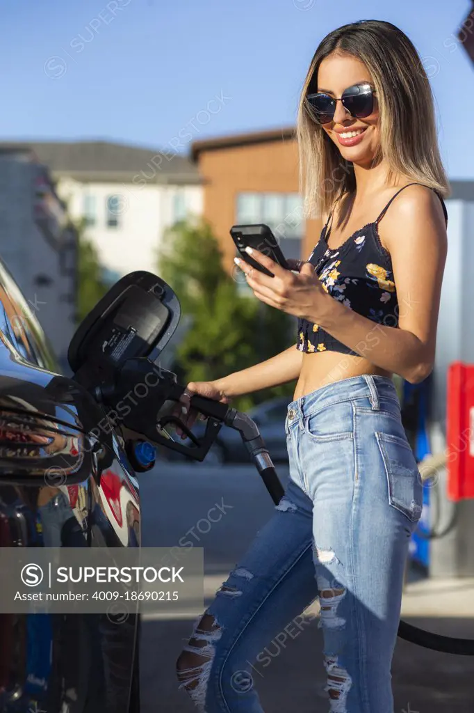 Young happy woman looking at mobile phone while pumping gas into car.