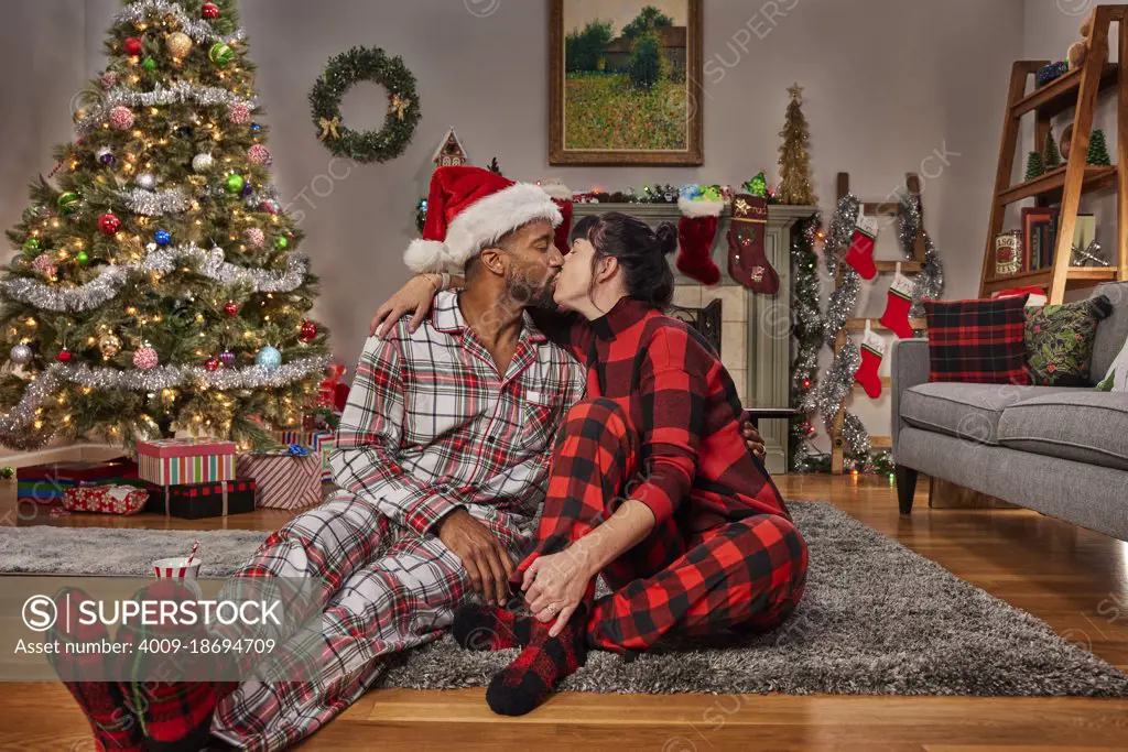 Romantic young diverse couple kissing while sitting in living room by Christmas tree.