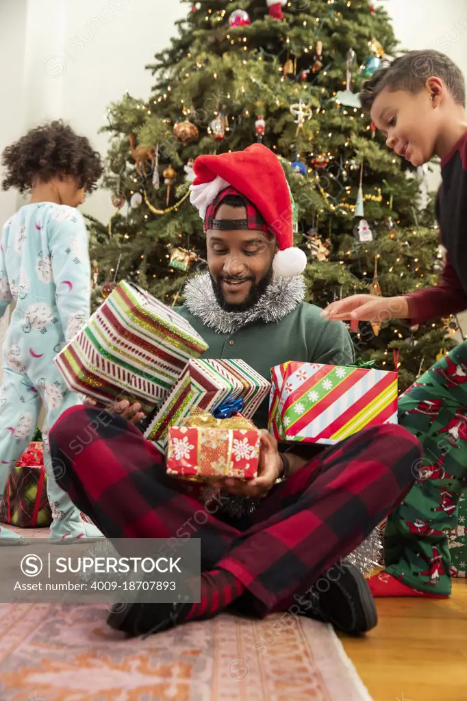 A father playing with his kids on Christmas Eve as they pile presents on his lap.