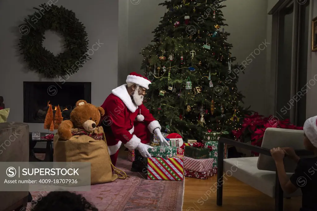 Black Santa Claus dropping off presents under Christmas tree in dark room, with a little girl peeking and watching behind chair unannounced. 