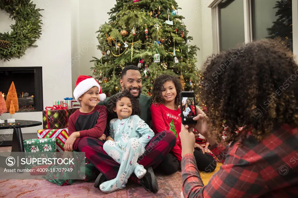 Young Black family taking pictures on mobile phone under Christmas tree.