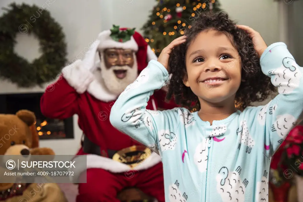 Young child in living room with a Christmas background and Santa mimicking her moves behind her. Black Santa.