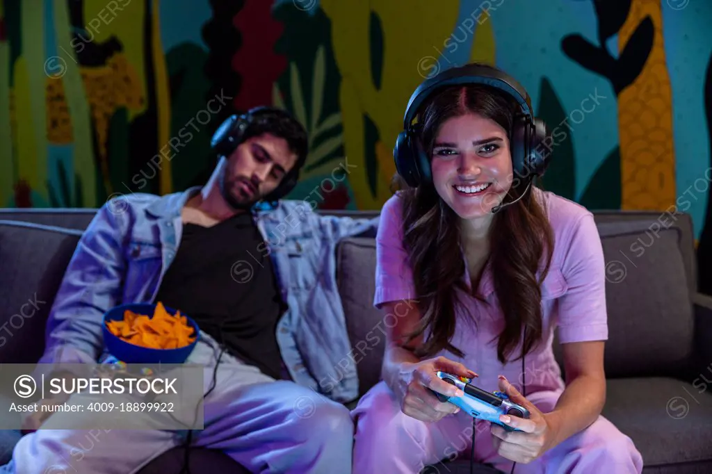 Smiling woman playing video games at night while her partner sleeps in background. 