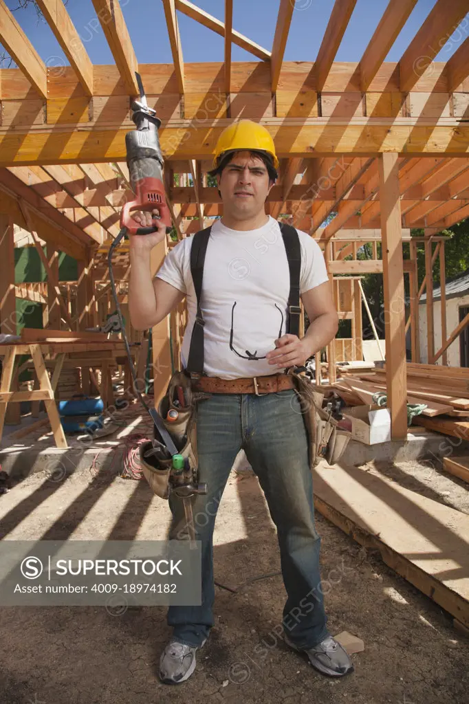 Construction worker wearing a hard hat, tool belt and holding an electric saw while standing on the construction site
