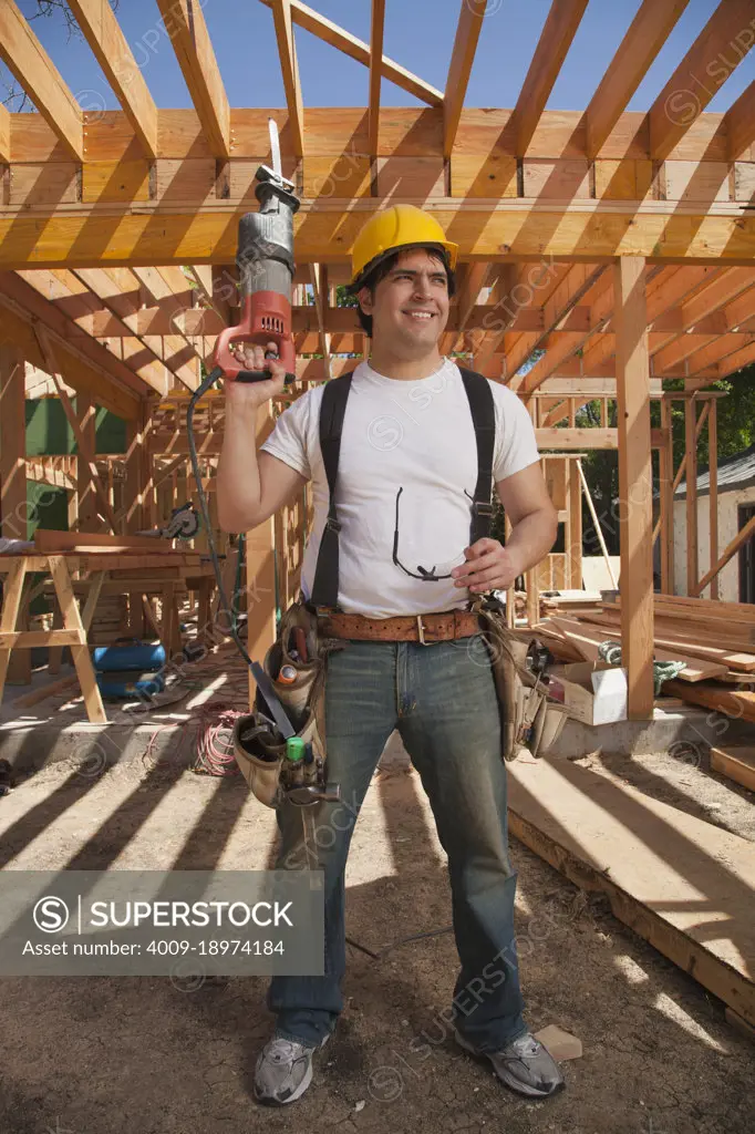 Construction worker wearing a hard hat, tool belt and holding an electric saw while standing on the construction site