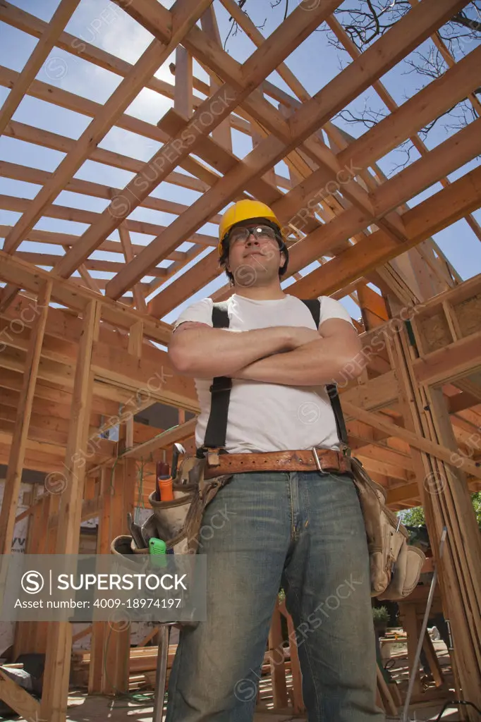 Portrait of a man wearing a hard hat and a tool belt with his arms crossed standing at a residential construction site