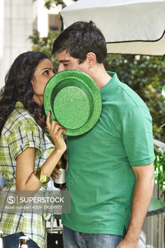 Young diverse couple kissing behind a green St. Patrick's Day hat while at party on balcony. 