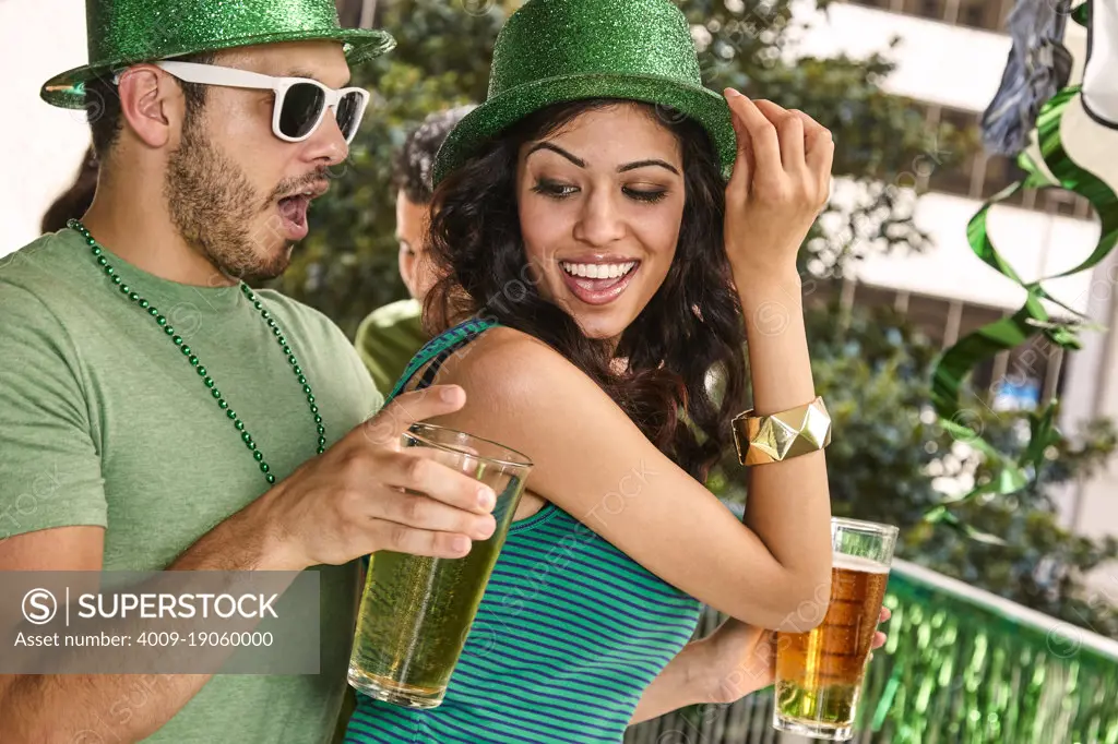 Young diverse couple dancing together wearing green sparkle hats and holding beer glasses for St. Patrick's Day.